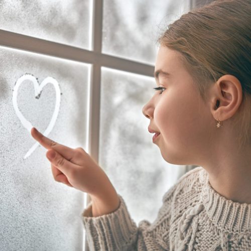 Cute little girl sitting by the window and drawing heart on frozen glass. Kid enjoys the winter.